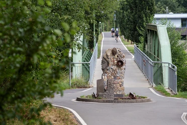 Steinskulptur „Zeitschienen“ auf einer früheren Bahntrasse vor grüner Bogenbrücke