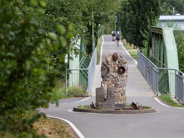 Steinskulptur „Zeitschienen“ auf einer früheren Bahntrasse vor grüner Bogenbrücke