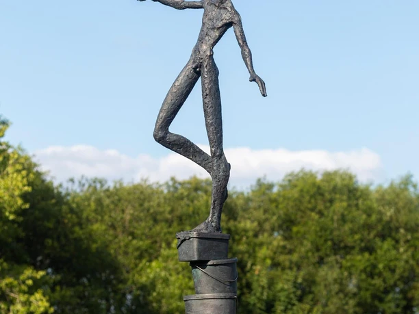 Bronzefigur balanciert auf gestapelten Eimern vor Hecken und blauem Himmel