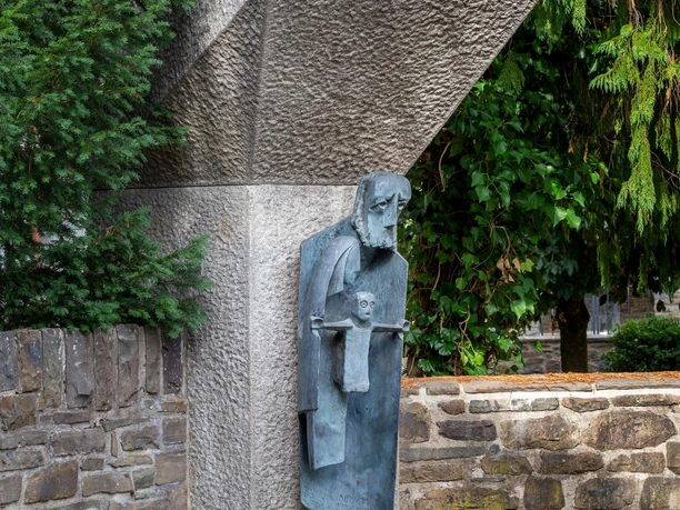 Steinskulptur Klausner Heinrich mit Bronzefigur vor Mauer und dichtem Grün