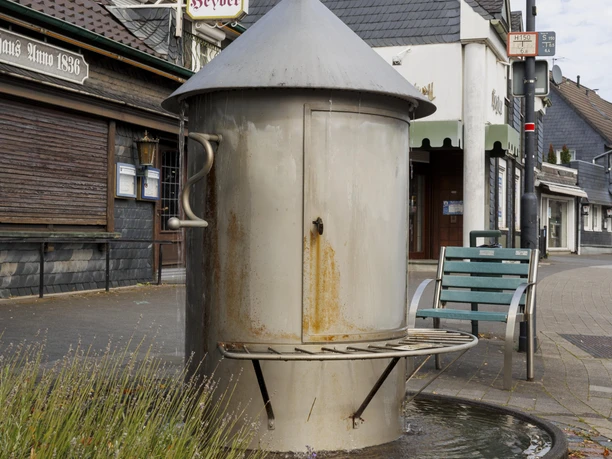 Metallener Dorfbrunnen in Burscheid-Hilgen vor Schieferhäusern und Sitzbank