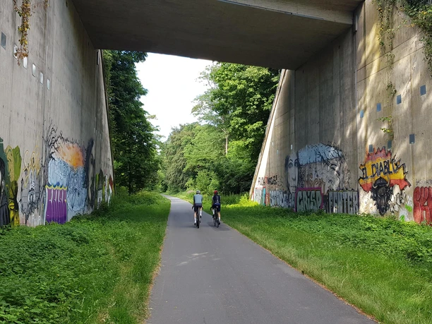 Graffiti an einer Brückenunterführung, zwei Radfahrer auf grünem Trassenweg