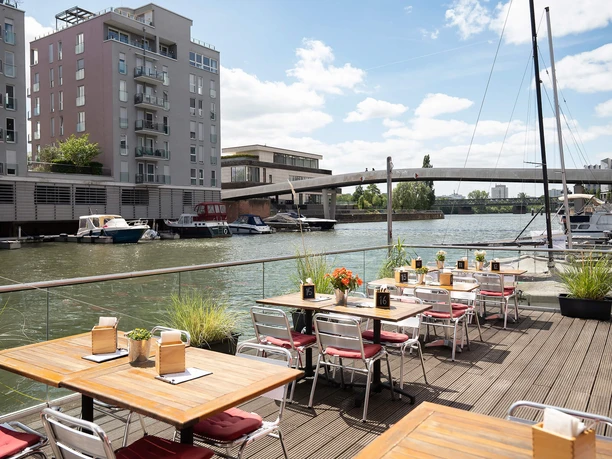 Sonnige Terrasse der Kombüse am Main mit Holztischen, Booten und Blick auf eine Brücke