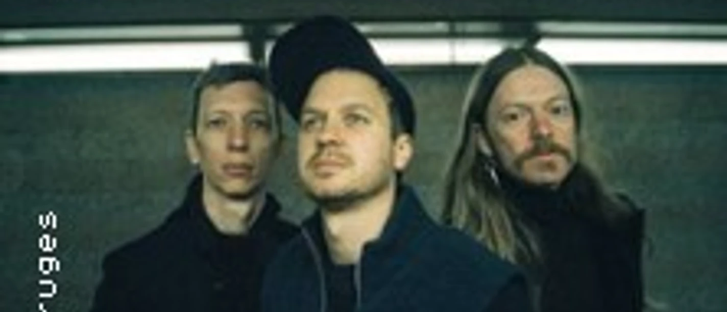 Drei Musiker von Bobby Rausch in dunkler Kleidung vor urbanem Hintergrund