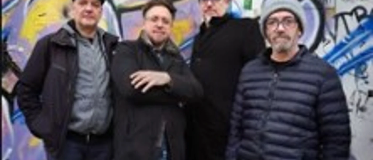 Vier Musiker posieren vor einer Graffitiwand, winterlich gekleidet.