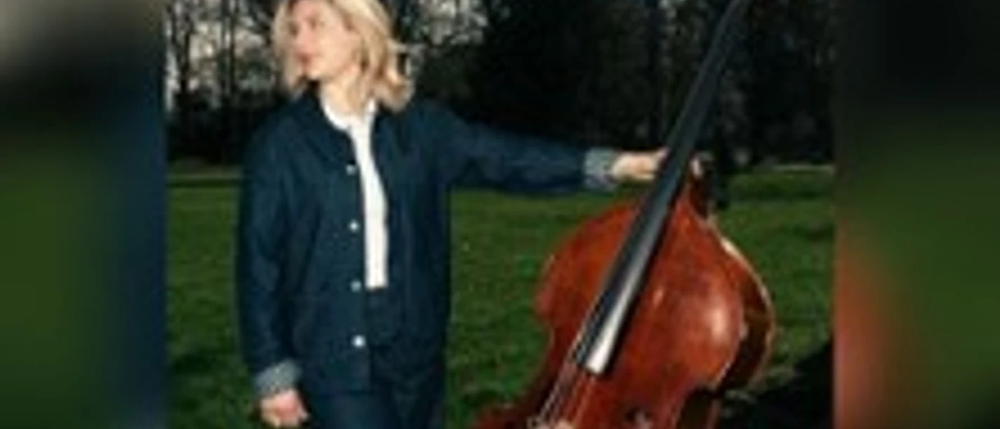 Blonde Musikerin steht im Park neben einem Kontrabass vor dunklen Bäumen.