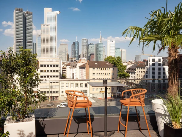 Rooftop-Bar mit Skylineblick auf Frankfurts Hochhäuser, Palmen und zwei orangefarbene Barstühle