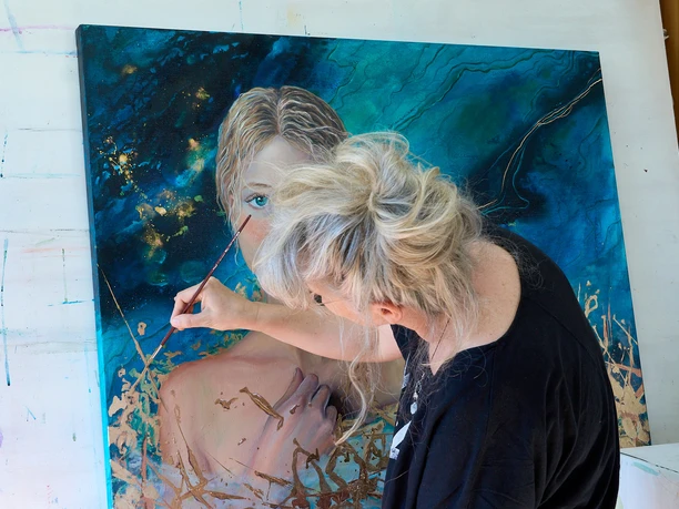 Künstlerin malt ein Porträt einer Frau mit blonden Haaren vor einem blauen Hintergrund.