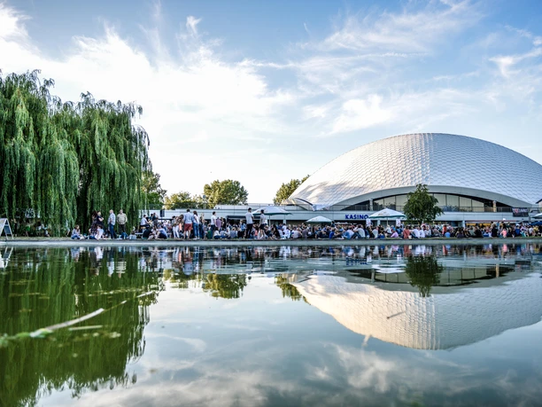 myticket Jahrhunderthalle Frankfurt_Kuppelförmiges Gebäude Kuppelförmiges Gebäude am Wasser mit Trauerweide und Menschen davor.