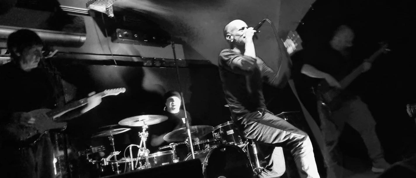 Schwarz-Weiß-Aufnahme einer Rockband mit Sänger, Gitarren und Schlagzeug auf dunkler Bühne