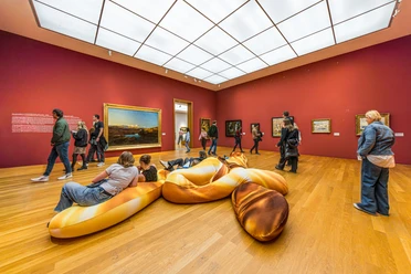 Ausstellungsraum des Museums der bildenden Künste mit roten Wänden und einer großen, goldfarbenen Liegemöglichkeit für Besucherinnen und Besucher im Zentrum.