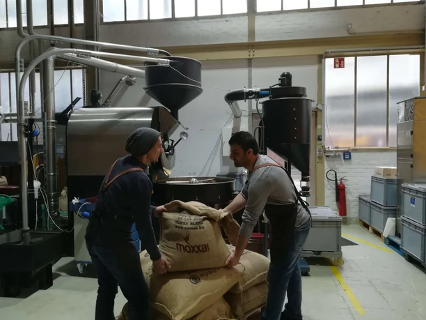 Zwei Personen heben einen Kaffeesack vor großen Röstanlagen.