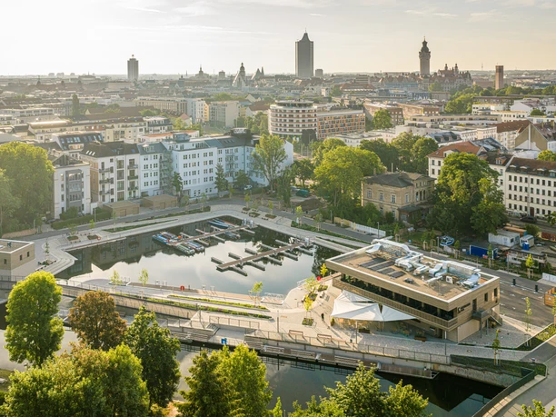 Luftaufnahme des Stadthafens Leipzig mit Bootsanlegern, Gastronomie und Skyline der Stadt im Hintergrund.