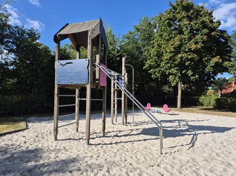 Klettergerüst mit Leitern und Wipptieren auf einem Sandspielplatz unter Bäumen.