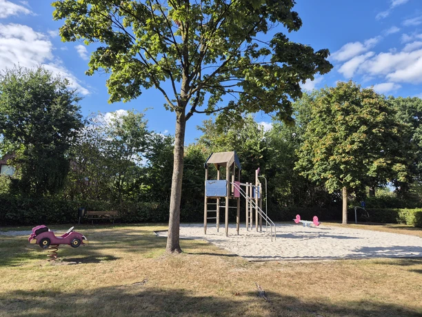 Spielplatz mit Kletterturm, Wipptieren und Sandfläche unter Bäumen.