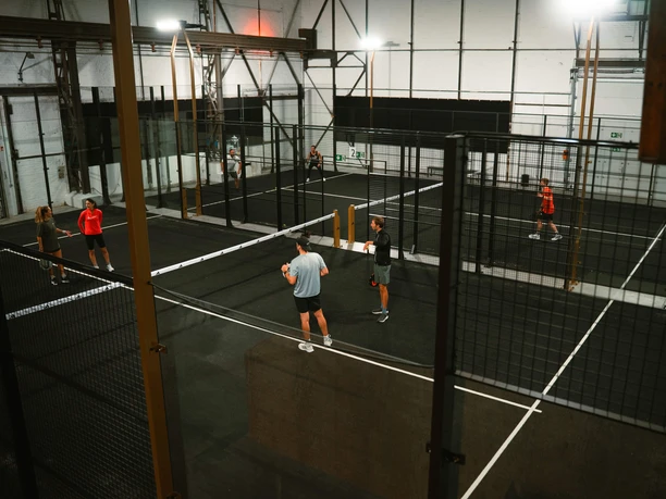 Mehrere Personen spielen Padel auf schwarzen Indoor-Courts in einer hohen Halle.