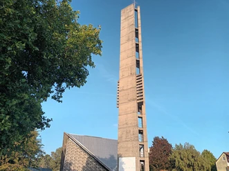 Betonturm der Kirche Heilige Dreifaltigkeit unter blauem Himmel, umgeben von Bäumen.