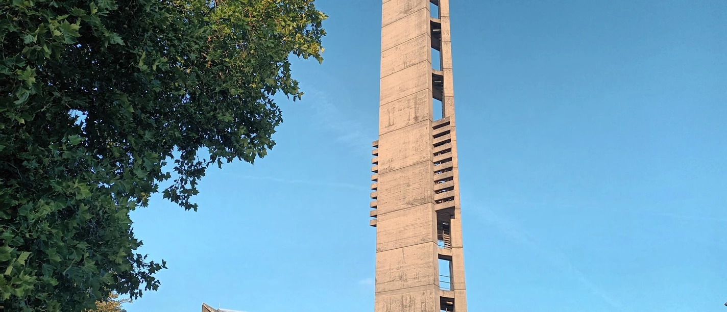 Betonturm der Kirche Heilige Dreifaltigkeit unter blauem Himmel, umgeben von Bäumen.