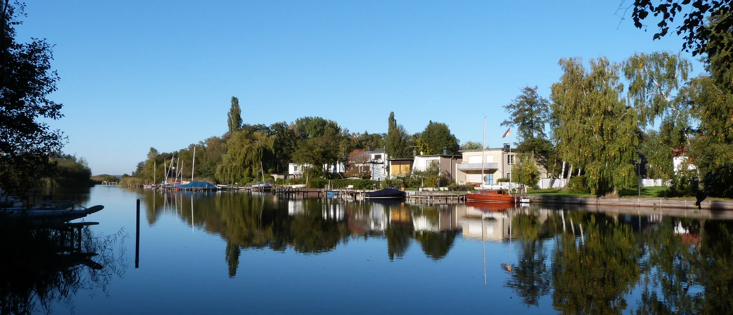 Grachten in Großenheidorn Canal with calm water, surrounded by trees and green houses, under a clear blue sky.