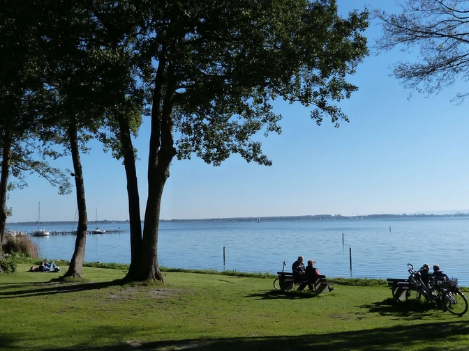 Mardorf Lüttjen Mardorf Ein ruhiger See mit Radfahrern und Spaziergängern auf einer Wiese unter Bäumen im Sonnenlicht.