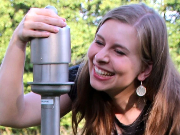 Wer der Sinne - Duftsäulen bei der Liebesinsel Frau lächelnd an Duftsäule auf Wiese, umgeben von grünem Wald. Emojis umfassen Freude und Natur.