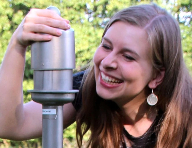 Wer der Sinne - Duftsäulen bei der Liebesinsel Frau lächelnd an Duftsäule auf Wiese, umgeben von grünem Wald. Emojis umfassen Freude und Natur.