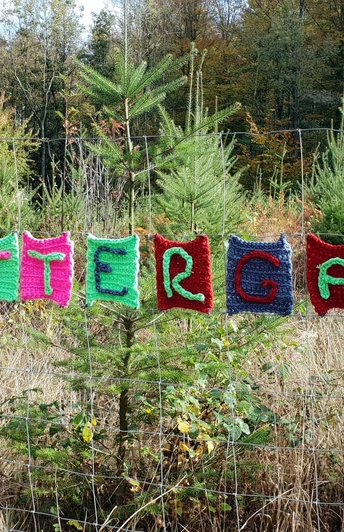 Bunte Strickbuchstaben formen das Wort "Schnattergang" an einem Maschendrahtzaun im Wald.