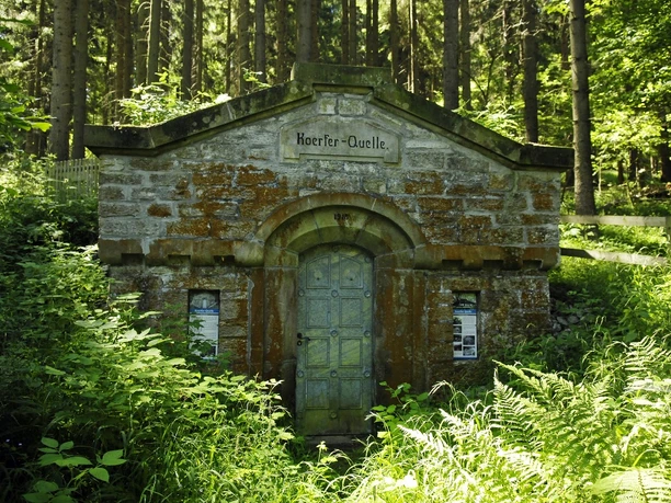 Historischer Quellhäuschenbau von Bäumen umgeben; beschriftete Tür und dichter Pflanzenbewuchs.