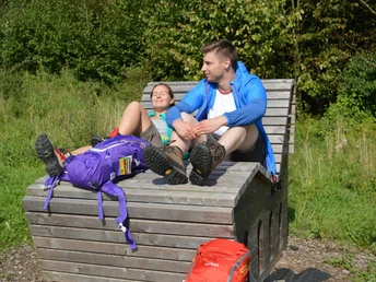 Zwei Personen entspannen auf einer Holzbank im Grünen nahe einem Wanderweg, umgeben von Bäumen.