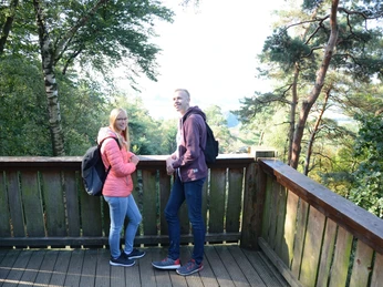 Zwei Personen stehen auf einer hölzernen Plattform in einem Wald und blicken in die Ferne.