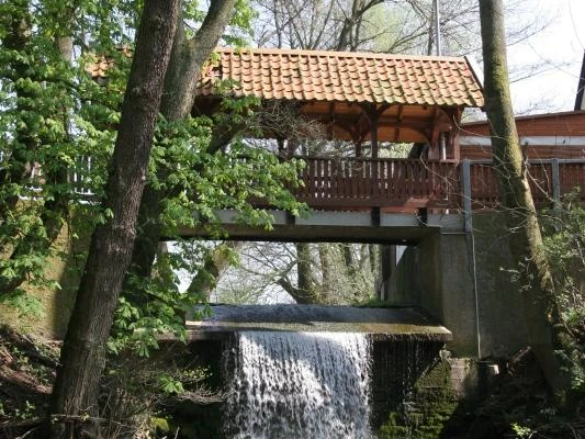 Ein kleiner Wasserfall fließt unter einer hölzernen Brücke hindurch, umgeben von Bäumen.