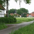 Anfahrt auf Bockhorst Ländliche Szene mit einer schmalen Straße durch grüne Felder und traditionellen Fachwerkhäusern.