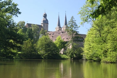 Abteikirche Blick auf die Abteikirche mit zwei spitzen Türmen, umgeben von grünen Bäumen und einem ruhigen See.