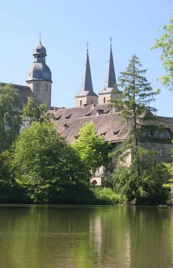 Abteikirche Blick auf die Abteikirche mit zwei spitzen Türmen, umgeben von grünen Bäumen und einem ruhigen See.
