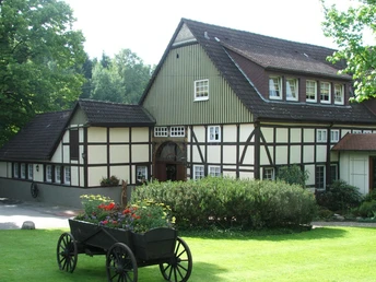 Fachwerkhaus mit Satteldach, umgeben von grünem Garten und Blumenkarren im Vordergrund.