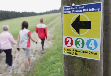 Nordic Walking auf dem Gesundheits- und Fitness-Parcours Bad Driburg 4 Drei Personen beim Nordic Walking auf einem Waldweg, ein Wegweiser mit Entfernungsangaben im Vordergrund.