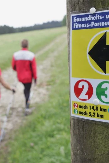 Nordic Walking auf dem Gesundheits- und Fitness-Parcours Bad Driburg 4 Drei Personen beim Nordic Walking auf einem Waldweg, ein Wegweiser mit Entfernungsangaben im Vordergrund.