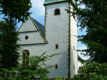 Historische Kirche mit grünem Turm und Uhr, umgeben von Bäumen und bunten Blumenbeeten.