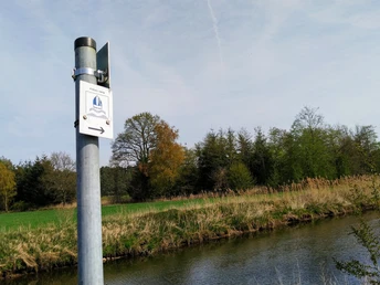 Wanderweg-Schild an einem Pfosten vor einer ruhigen Flusslandschaft mit Bäumen im Hintergrund.