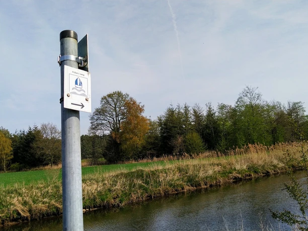 Prälatenweg Wanderweg-Schild an einem Pfosten vor einer ruhigen Flusslandschaft mit Bäumen im Hintergrund.