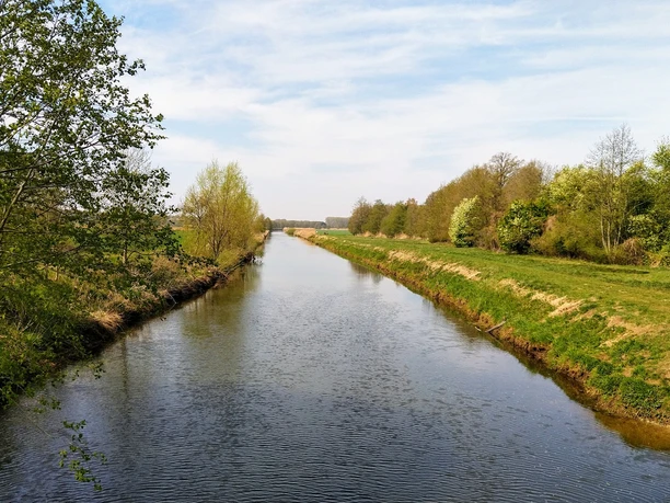 Ems - Prälatenweg Ein ruhiger Fluss in einer von Bäumen gesäumten ländlichen Landschaft unter klarem Himmel.