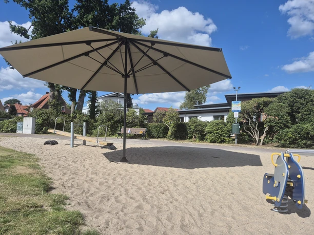 Sandspielplatz mit großem Sonnenschirm, Wippe und Federwipptier unter blauem Himmel.