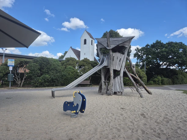 Spielplatz mit Holzturm, Rutsche und Wipptier auf Sandfläche vor einer Kirche.
