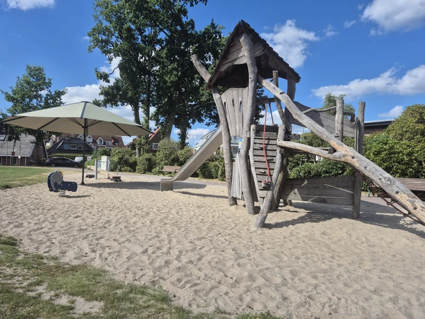 Sandspielplatz in Rastede mit Holz-Kletterturm, Rutsche, Wippe und Sonnenschirm.