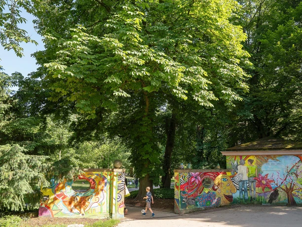 Bunter Parkeingang mit Graffiti und Kind in den Günthersburgpark unter grünen Bäumen