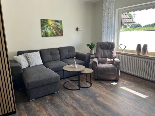 Wohnzimmer mit grauem Ecksofa, Sessel, runden Tischen und großem Fenster.