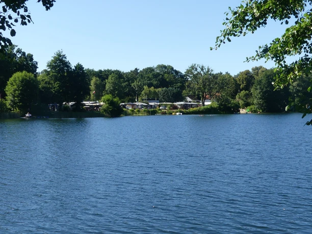 Waldsee - im Hintergrund der Campingplatz Ein ruhiger See mit blauem Wasser, umrahmt von Bäumen. Im Hintergrund ein Campingplatz sichtbar.