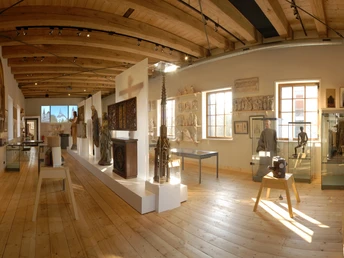 Ein heller Museumsraum mit Holzdecke, Exponaten in Vitrinen und Skulpturen auf einem Podest.