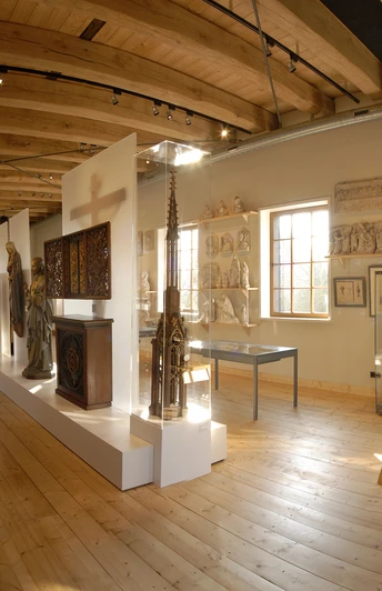 Ein heller Museumsraum mit Holzdecke, Exponaten in Vitrinen und Skulpturen auf einem Podest.