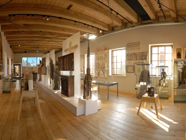 Ein heller Museumsraum mit Holzdecke, Exponaten in Vitrinen und Skulpturen auf einem Podest.